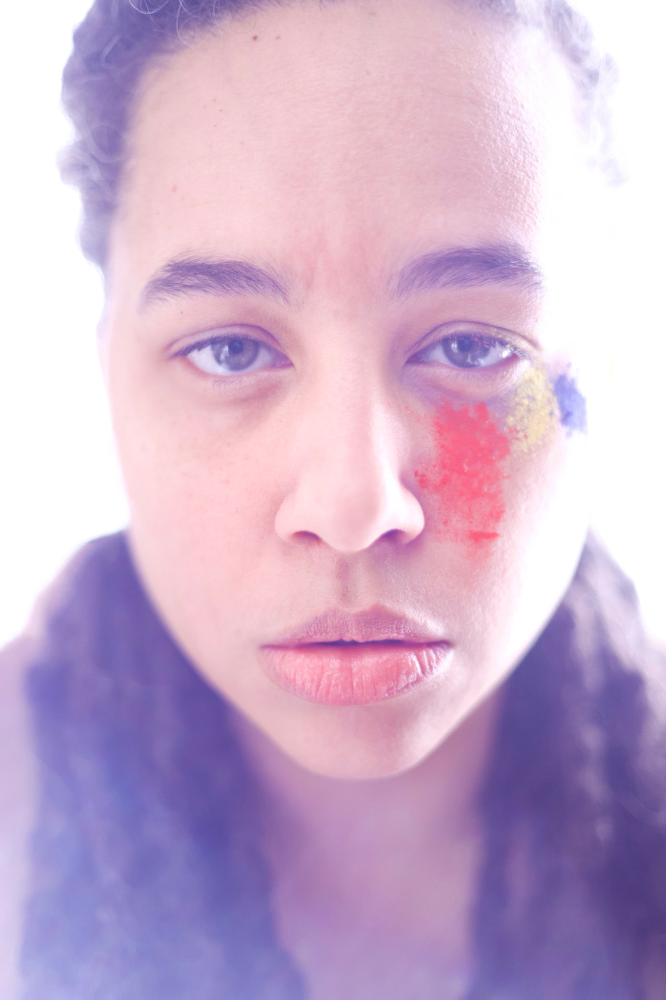 Closeup backlit headshot of a person staring into camera with colored powder beneath their left eye in red, yellow, and blue