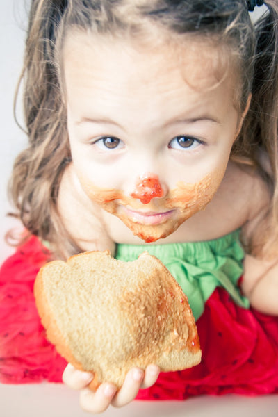 Child wearing a strawberry dress, looking up with peanut butter and jelly smeared on their face and a slice of bread they are holding