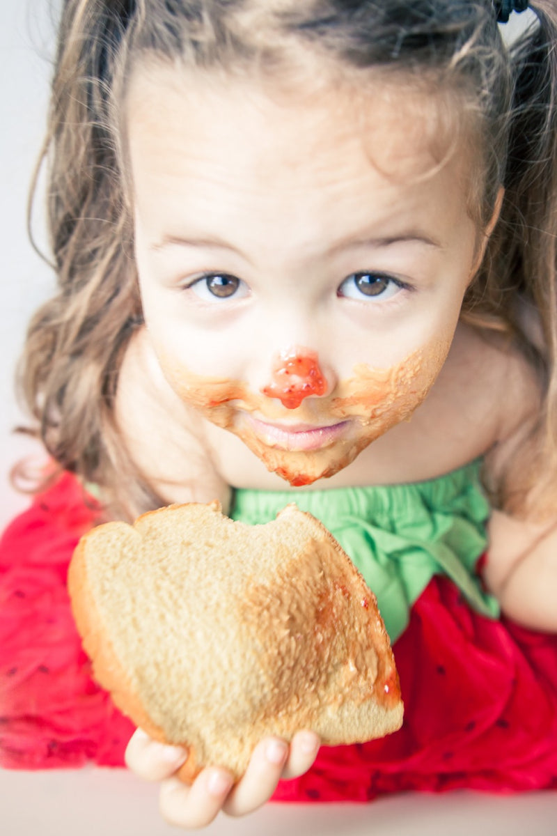 Child wearing a strawberry dress, looking up with peanut butter and jelly smeared on their face and a slice of bread they are holding