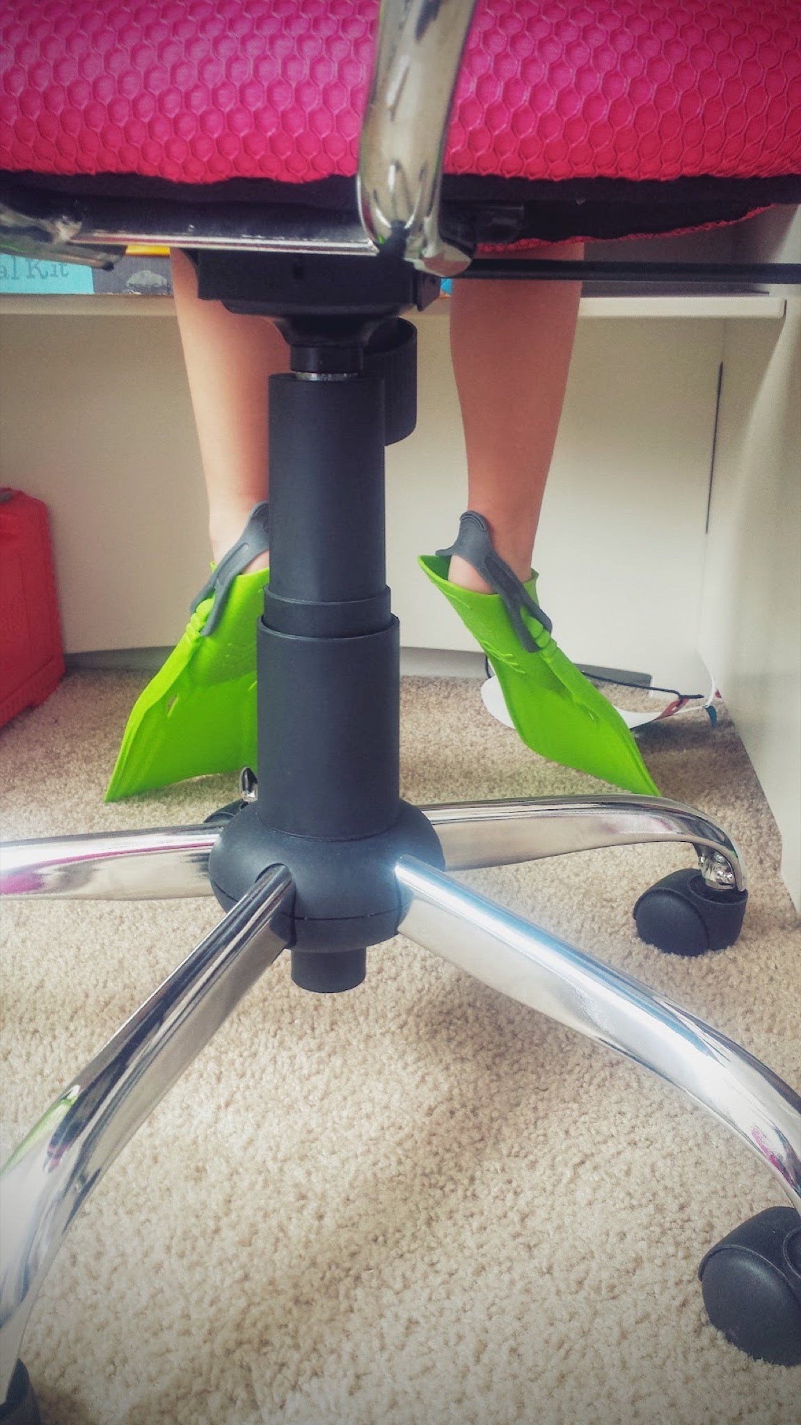 Closeup of a child's legs in neon green flippers, sitting at a white desk in a neon pink computer chair on white carpet