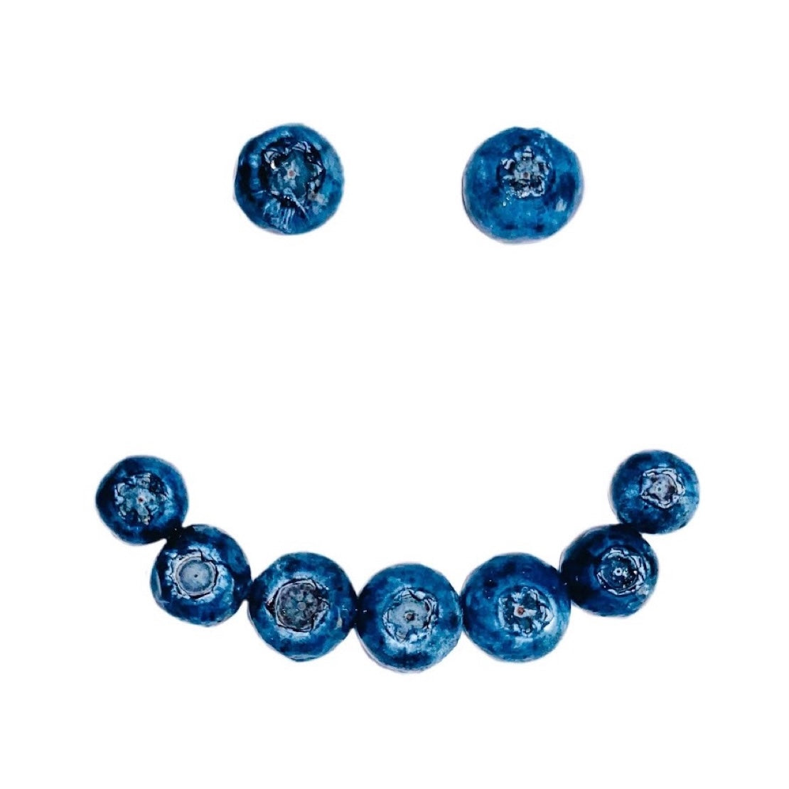 Blueberries arranged on a white background into a smiley face with a single blueberry for each eye and multiple for the smiling mouth