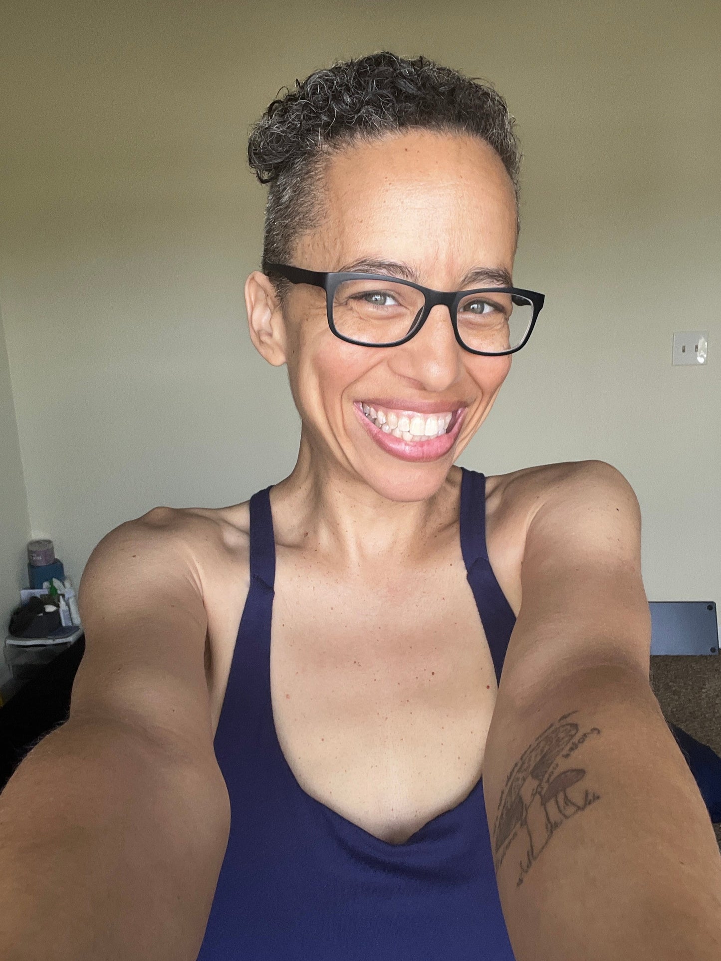 Blue Dulac smiling big, wearing a blue jumpsuit, holding the phone with both hands for a selfie with her mushroom tattoo on left arm visible