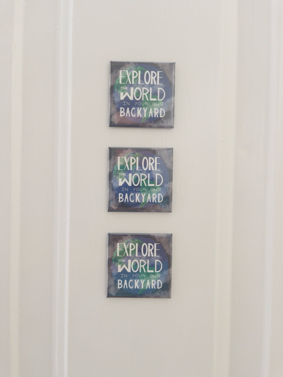 Three identical decorative magnets with 'EXPLORE THE WORLD IN YOUR OWN BACKYARD' text on a white door